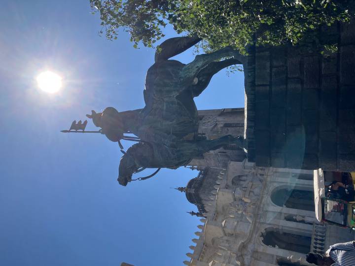 Silhouette of an equestrian statue standing before an old stone cathedral beneath a bright blue sky and sun.