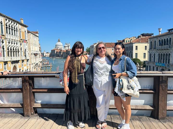 Three travellers on a wooden bridge over the Grand Canal with Santa Maria della Salute and Venetian palaces behind.