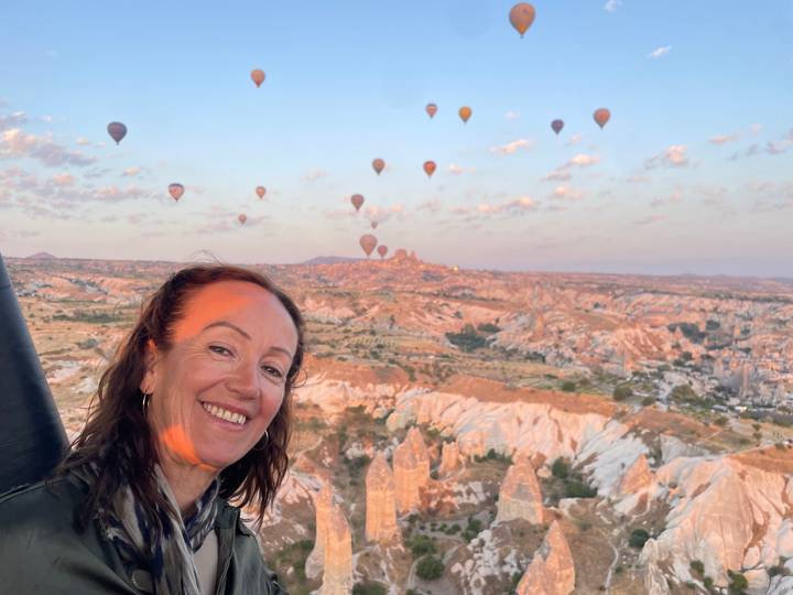 Smiling woman in hot-air balloon basket with dozens of colorful balloons floating over Cappadocia landscapes