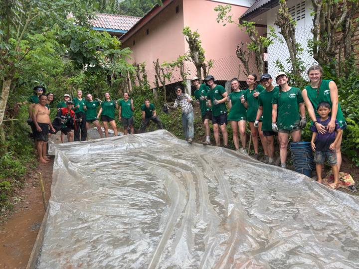 Large group of young volunteers in green shirts standing beside a long plastic sheet on a muddy jungle path.
