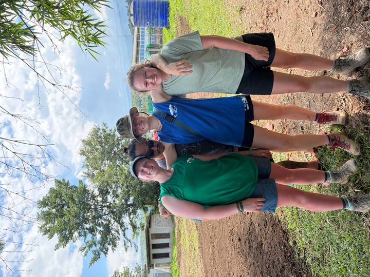 Four muddy volunteers in casual sportswear pose arm-in-arm on a rural worksite with trees and blue sky.