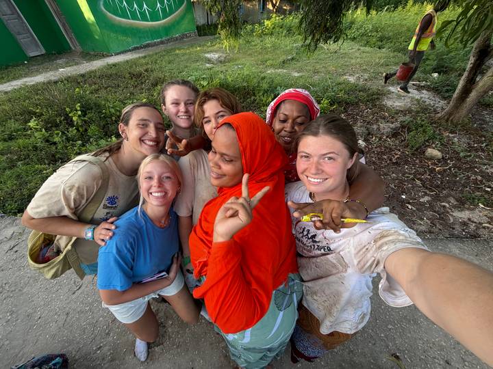 Smiling women crowd together for a cheerful selfie, one wearing a bright red scarf, outdoors on a grassy roadside.