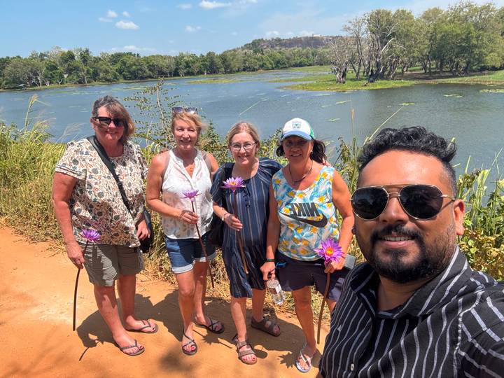 Tour guide and four women holding lotus flowers smile for a selfie beside a tranquil lake with lily pads.
