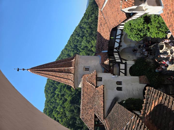 Medieval courtyard with red-tiled tower and forested mountain backdrop, filled with tourists exploring.