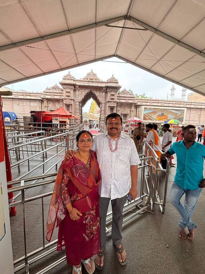Smiling couple with tilak marks pose at the entrance of a Hindu temple complex with metal queue rails.
