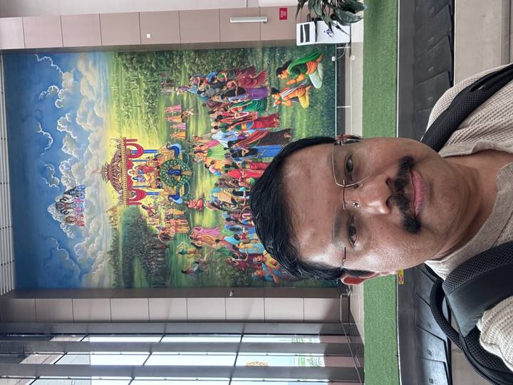 Man takes a selfie in front of a large colourful epic Hindu mural inside an airport terminal.