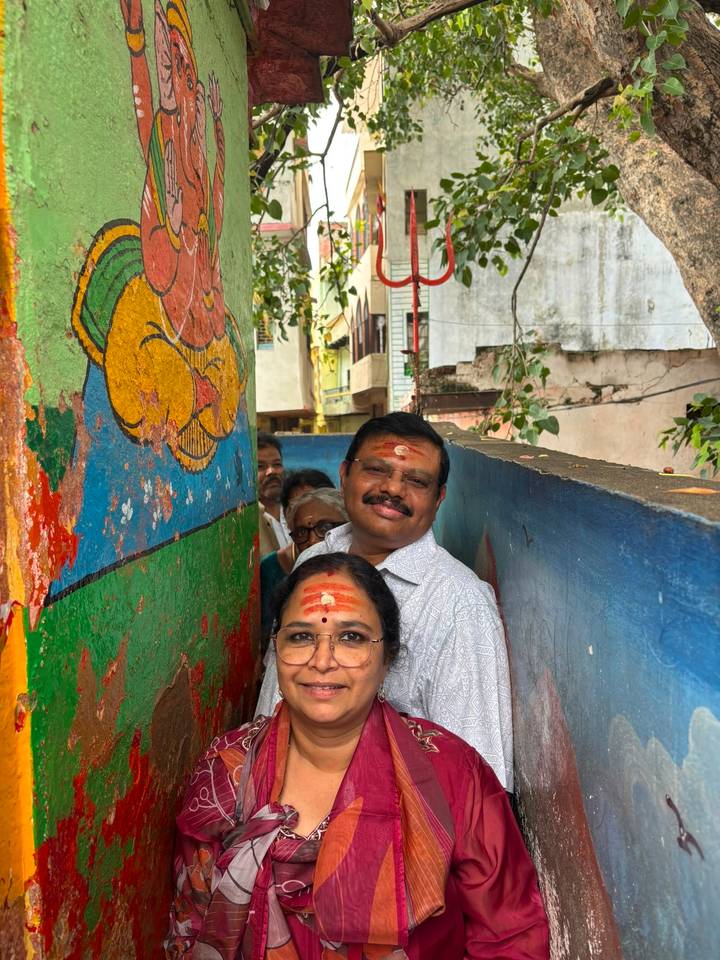 Couple with vermilion marks stand in a narrow alley painted with bright Hindu motifs and trees overhead.