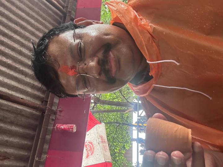 A middle-aged man wearing an orange rain poncho and tilak smiles while holding a small clay cup of tea inside a covered stall.
