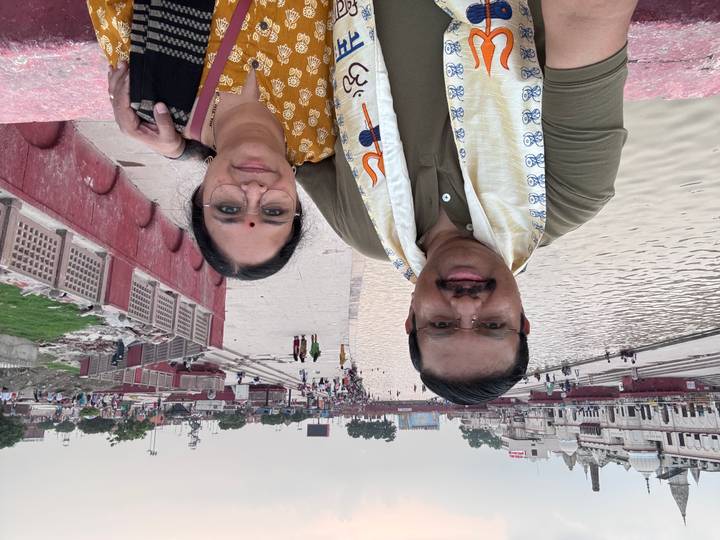 A couple takes a selfie on a broad riverside walkway with people strolling in the distance and water on the right.