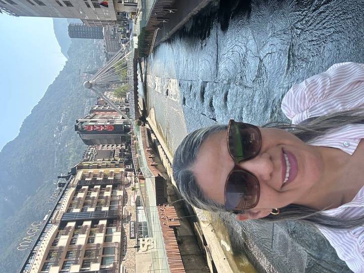 Woman taking a selfie beside a river and modern pedestrian bridge with mountains behind in Andorra La Vella