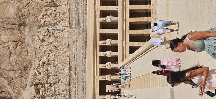 Visitors walk up the broad ramp toward the colonnaded facade of an ancient Egyptian temple carved into a cliff.