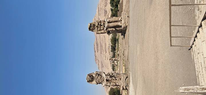 Two colossal seated statues rise from a barren plain with distant hills under a bright blue sky.