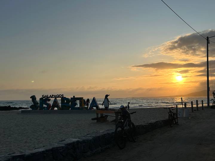 Beachfront sign reading 'Galapagos Isabela' silhouetted at sunset with ocean waves and a glowing sky.