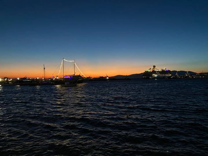 Colorful sunset over a harbor with sailboats and a lit cruise ship silhouetted against the horizon