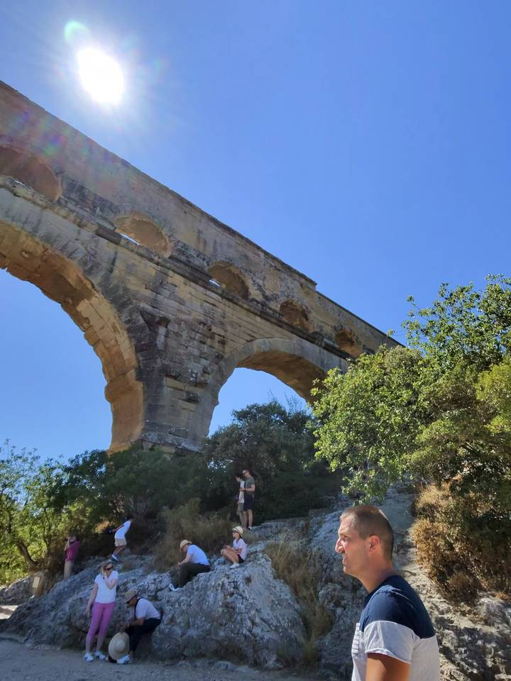 Stone arches of an ancient Roman aqueduct rise against a clear blue sky with surrounding greenery.
