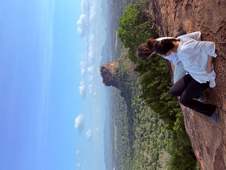 Two travelers relax on a rocky ledge overlooking the forested plain and iconic Sigiriya Rock rising in the distance.