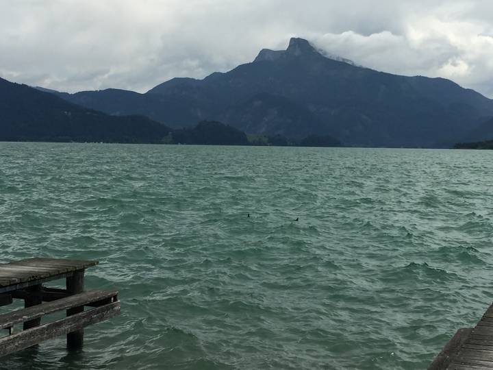 Choppy turquoise lake with wooden jetty and distant mountain backdrop under grey skies