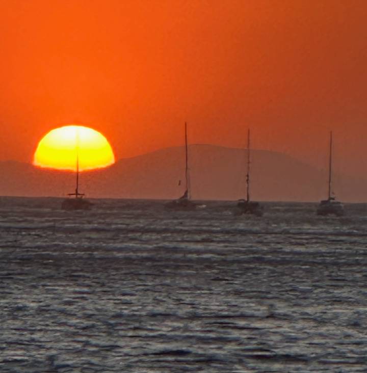 Brilliant orange sunset over the sea with silhouettes of several sailboats and a distant island.