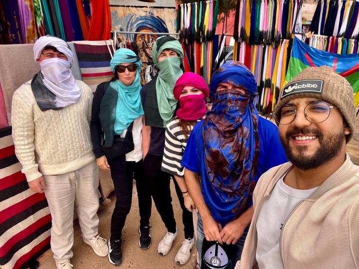 Travellers in colourful headscarves pose for a selfie amid fabric stalls in a Moroccan market.