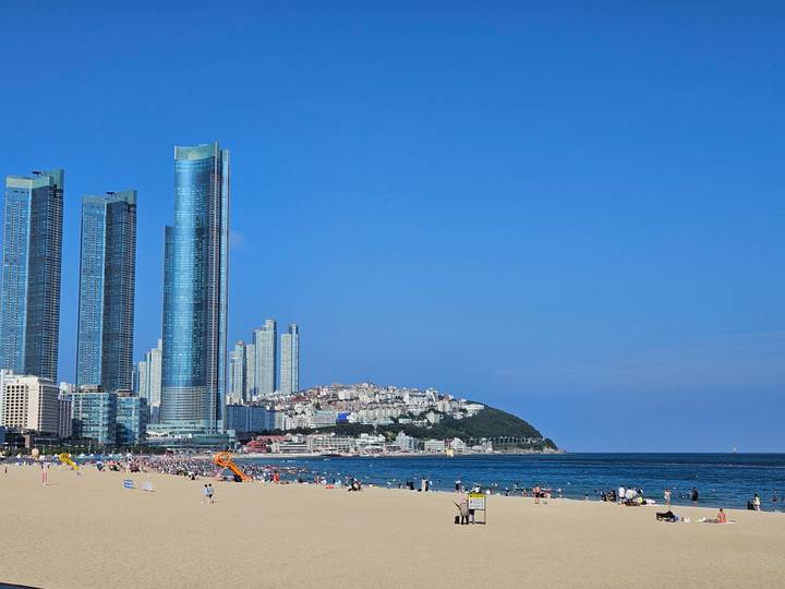 Busy beach scene with tall modern skyscrapers rising behind crowded sand and blue sea.