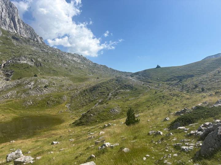 Grassy alpine hollow with a small pond nestles amid rocky slopes under a bright sky.