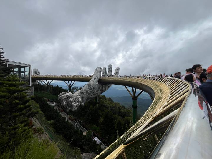 The curving Golden Bridge supported by giant stone hands stretches across misty mountains filled with tourists.