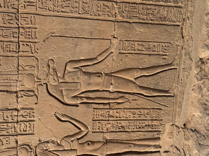 Close-up relief carving of an Egyptian god with hieroglyphics on a sandstone wall lit by warm sunlight.