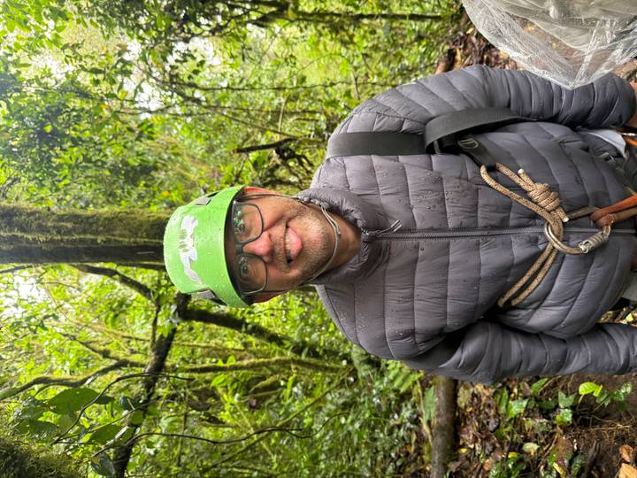 Portrait of a smiling man wearing a helmet and climbing harness in a misty green rainforest.