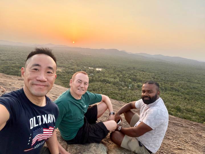 Three travelers resting on a rocky summit overlooking forested plain at sunset