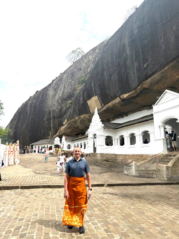 Visitor standing before white-washed monastery buildings built beneath an overhanging rock cliff