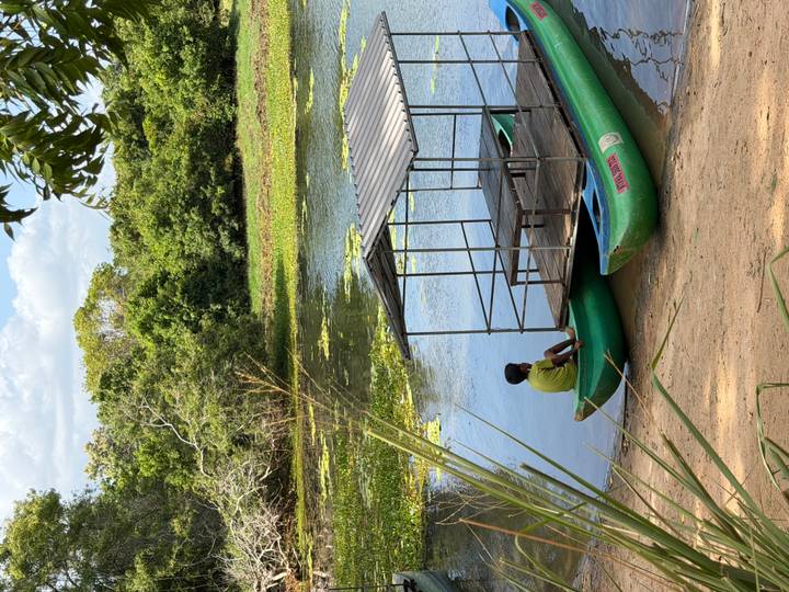 Young boy sits in a green canoe beside a simple metal-roofed raft on a tranquil, lily-covered river.