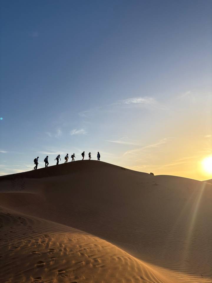 Silhouettes of hikers cresting a dune against a golden desert sunset.