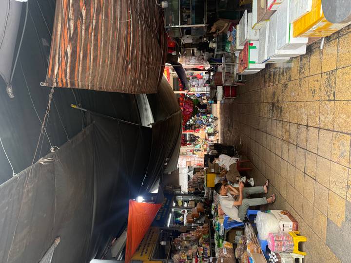 Covered wet market aisle with stalls, tarps overhead and locals sitting on plastic chairs.