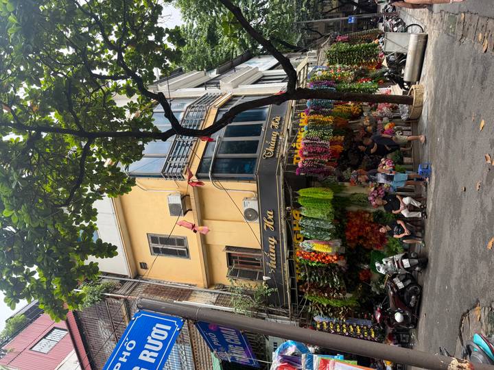 Corner flower shop in Hanoi with colorful artificial blooms hanging from awning and people seated below.