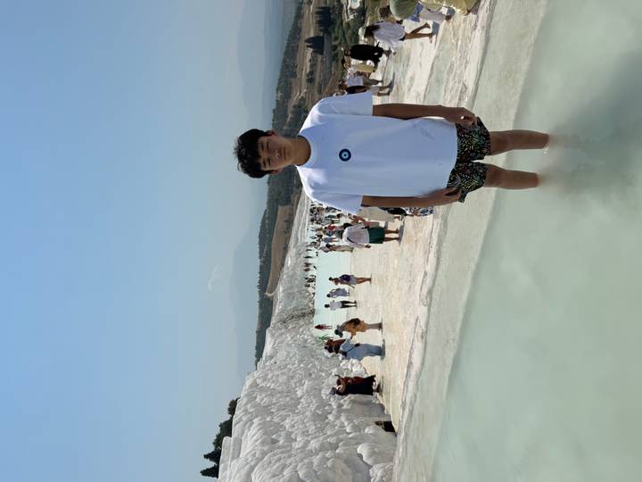 Teen boy posing barefoot in the shallow turquoise pools of Pamukkale with many tourists walking along the white calcium terraces in the background.
