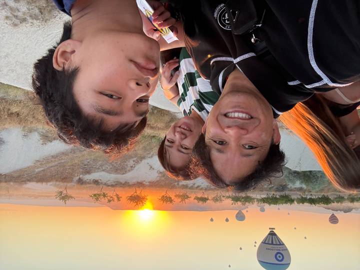Family selfie at sunrise with Cappadocia’s hot-air balloons rising above golden valleys and a glowing orange horizon.