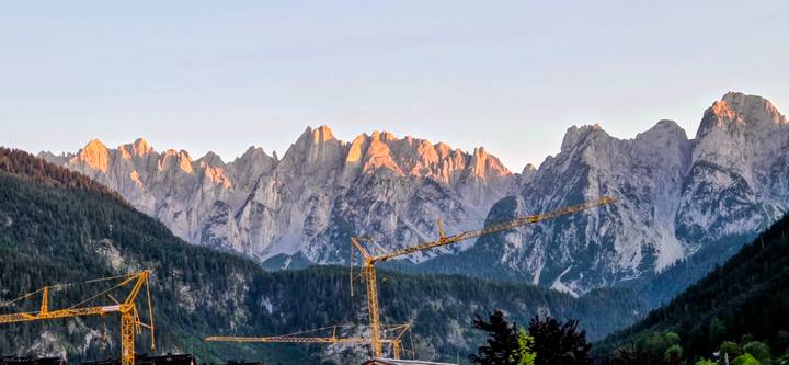 Construction cranes stand before rugged alpine peaks glowing in the warm light of sunset.