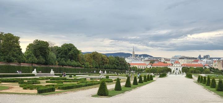 Gravel boulevard lined with cone-shaped shrubs leads through Belvedere Gardens toward Vienna’s skyline.