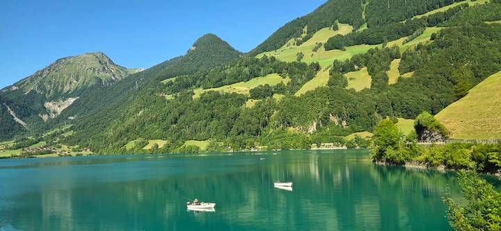 Rowboats drift on a crystal-clear turquoise alpine lake bordered by rolling green hills and forests.