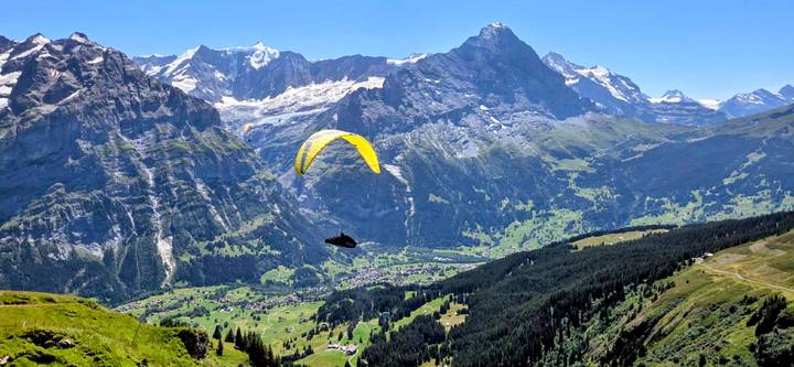 Bright yellow paraglider soars above a vast alpine valley backed by dramatic snowy peaks.