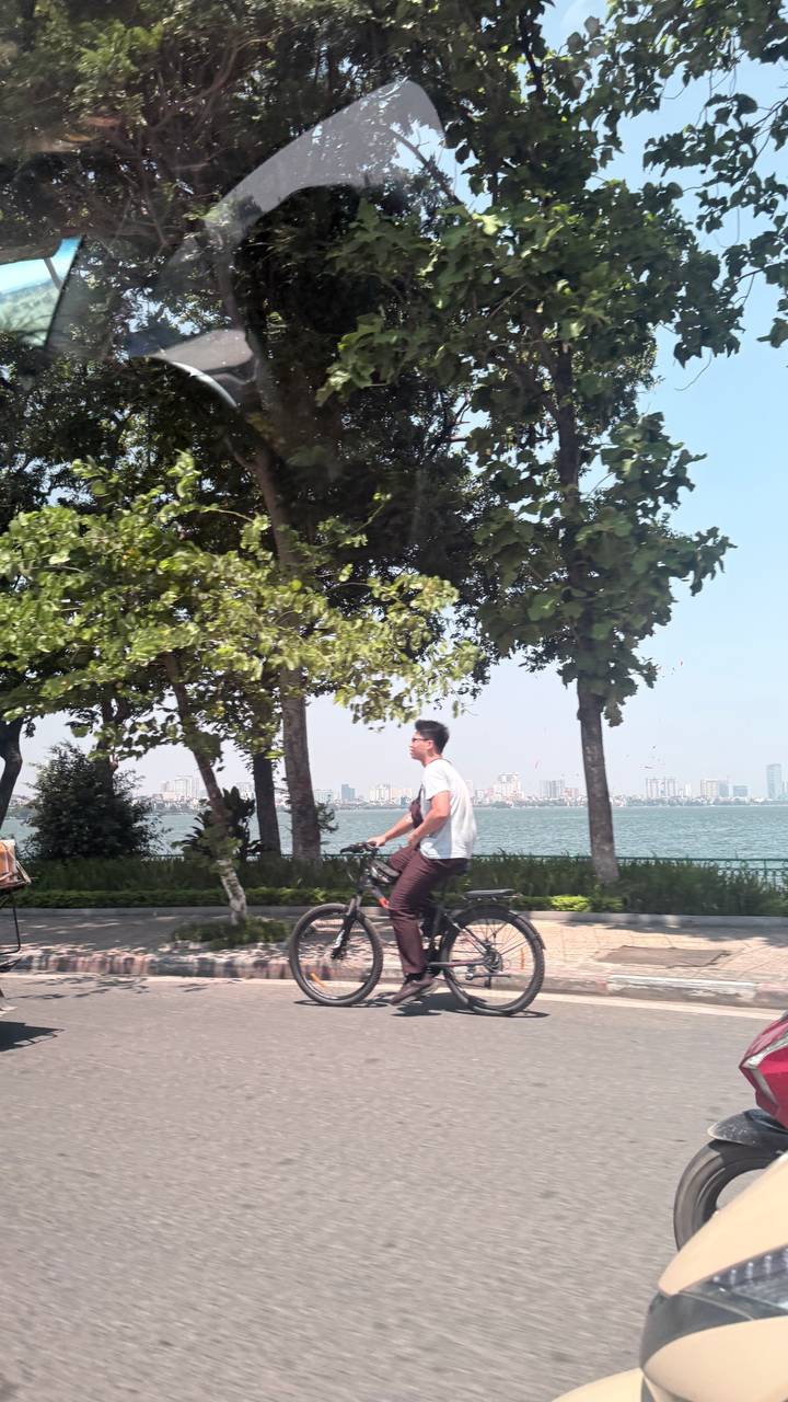 Man cycling along lakeside path with city skyline across the water, motion blur in foreground trees.