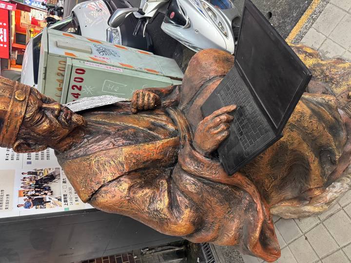 Bronze statue of ancient scholar seated and writing on a tablet in city street.