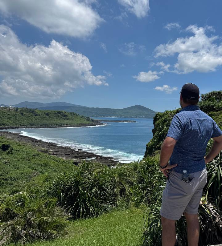 Traveler stands on cliffside overlooking blue sea and green hills of Kenting coast.