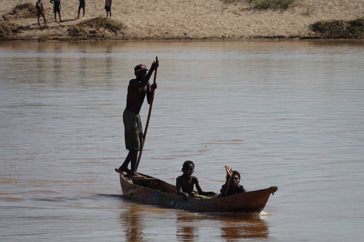 Traditional wooden canoe with three locals, one standing to pole, crossing a brown river.