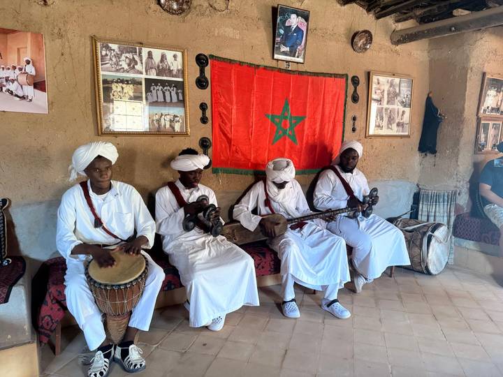 Four musicians in white traditional dress perform with drum and string instruments before a Moroccan flag inside an adobe room.