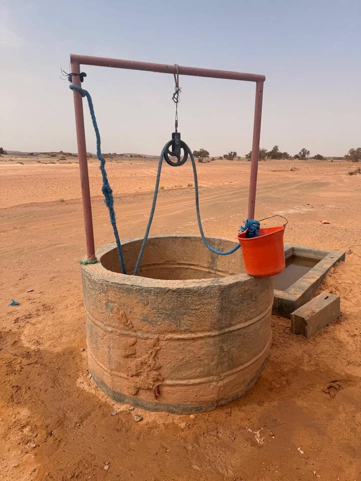 Simple desert well with rope, pulley and orange bucket stands in sandy landscape.