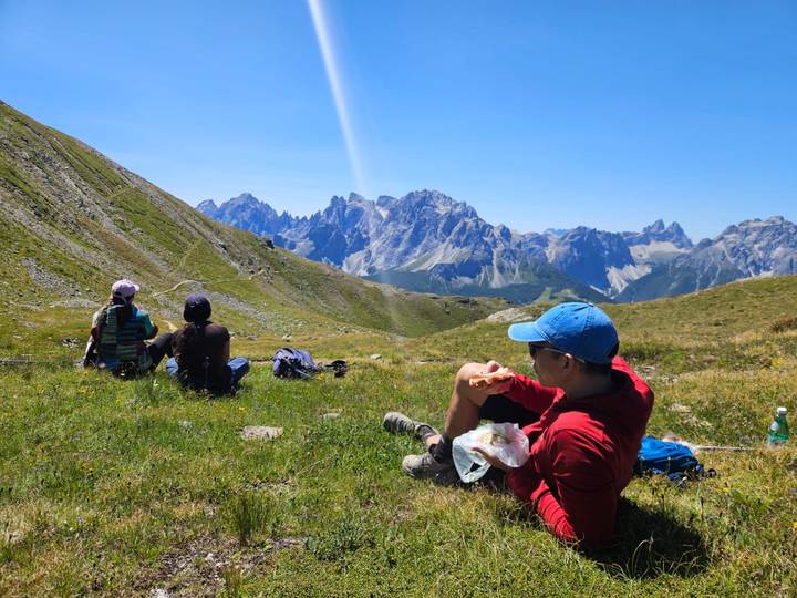 Hikers rest on an alpine meadow enjoying lunch while admiring jagged Dolomite peaks under a clear sky.