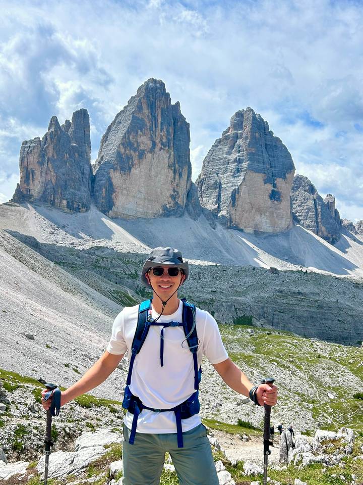 Hiker smiles in front of the dramatic vertical walls of Tre Cime on a sunny day.