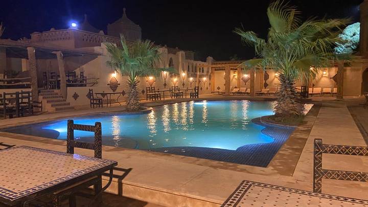 An atmospheric riad courtyard at night with a lit turquoise swimming pool and palm trees.