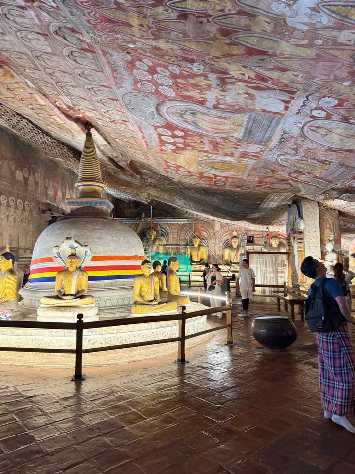 Ornate cave temple interior with golden Buddha statues and vivid painted ceiling, visitors exploring the hall